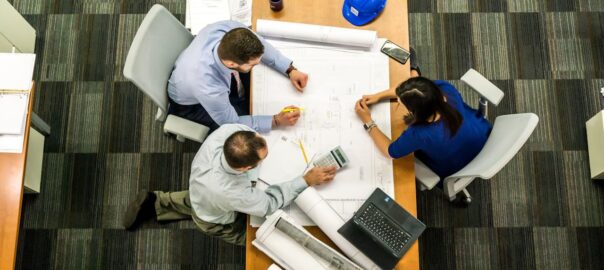 Three professionals reviewing architectural blueprints and plans at a conference table with a blue hard hat and laptop overhe