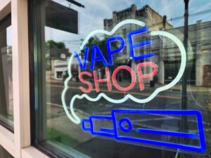 Neon vape shop sign in a storefront window displaying "VAPE SHOP" text with a glowing e-cigarette illustration